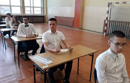 Zdjęcie maturzystów siedzących w przy szkolnych ławkach na sali gimnastycznej 