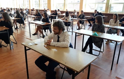 Zdjęcie maturzystów siedzących w przy szkolnych ławkach na sali gimnastycznej 