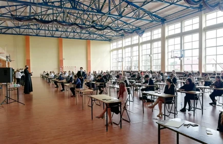 Zdjęcie maturzystów siedzących w przy szkolnych ławkach na sali gimnastycznej 