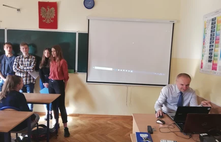 Na zdjęciu uczniowie ustawieni pod tablicą oraz siedzący nauczyciel