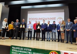 Zdjęcie przedstawia osoby stojące na scenie wraz z trofeami