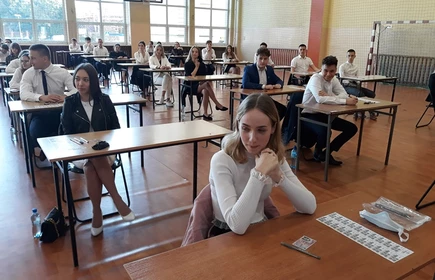 Zdjęcie maturzystów siedzących w przy szkolnych ławkach na sali gimnastycznej 