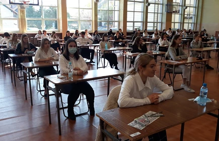 Zdjęcie maturzystów siedzących w przy szkolnych ławkach na sali gimnastycznej 